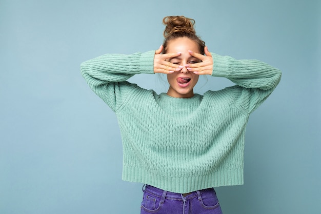 Photo photo of young positive happy joyful beautiful blonde lady with sincere emotions wearing casual blue pullover isolated over blue background with copy space and covering eyes with hands and having fun.