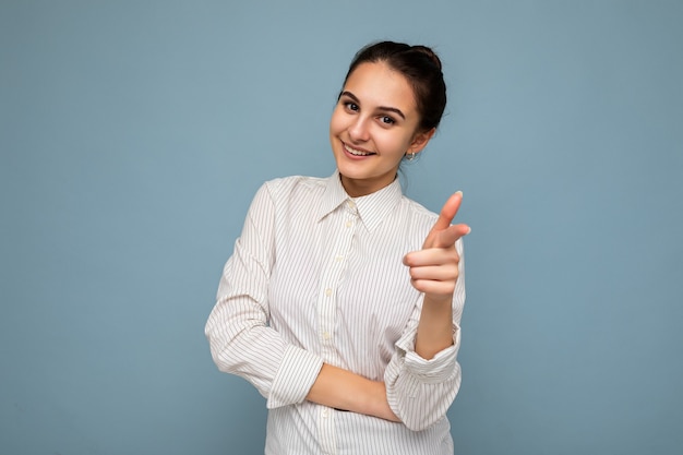 Photo of young positive happy smiling beautiful woman with sincere emotions wearing stylish clothes isolated over background with copy space and pointing at camera