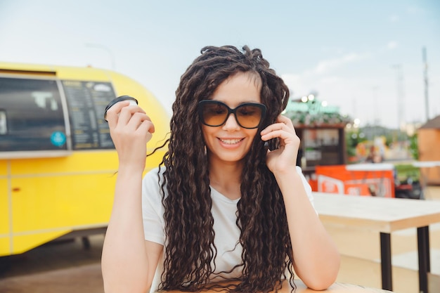 A photo of a young smiling happy woman who sits at the food court of a shopping center at a table and works at a computer laptop talks on a mobile phone Freelance concept