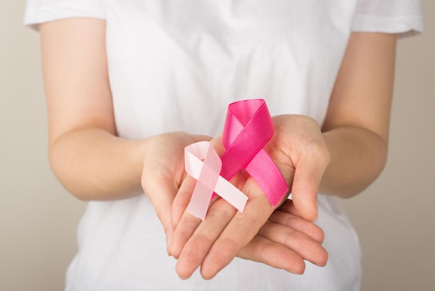 Photo of young woman in white tshirt holding two pink ribbons in palms symbol of breast cancer awareness on isolated grey background