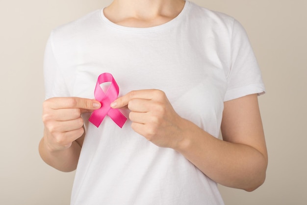 Photo of young woman in white tshirt holding vivid pink ribbon symbol of breast cancer awareness on isolated grey background