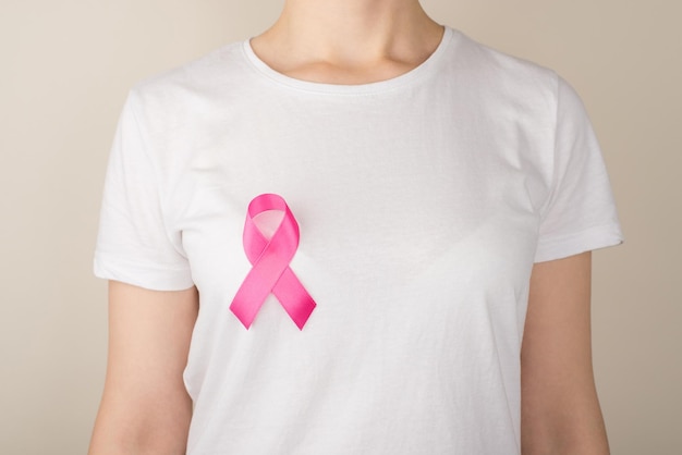 Photo of young woman in white tshirt with pink ribbon symbol of breast cancer awareness on isolated grey background