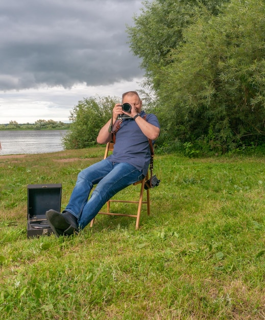 A photographer with a camera sits on the bank by the river in summer