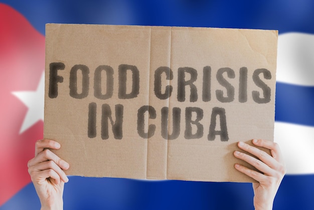 The phrase Food crisis in Cuba is on a banner in men's hands with a blurred Cuban flag in the background Crisis Finance Life Nutrition Bread Disaster Collapse Social issue