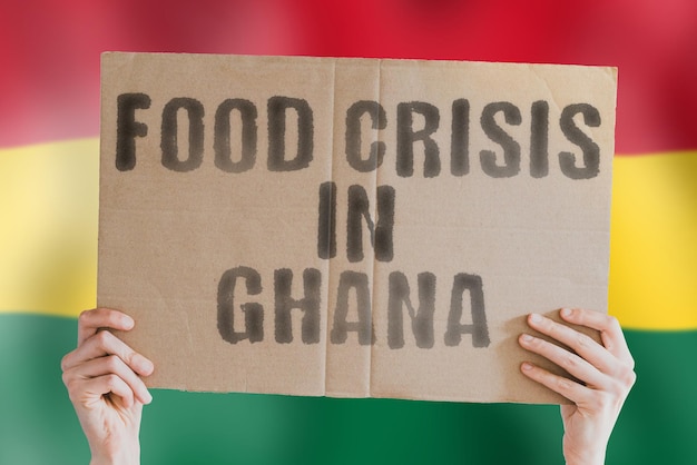 The phrase Food crisis in Ghana is on a banner in men's hands with a blurred Ghanaian flag in the background Crisis Finance Life Nutrition Bread Disaster Collapse Social issue