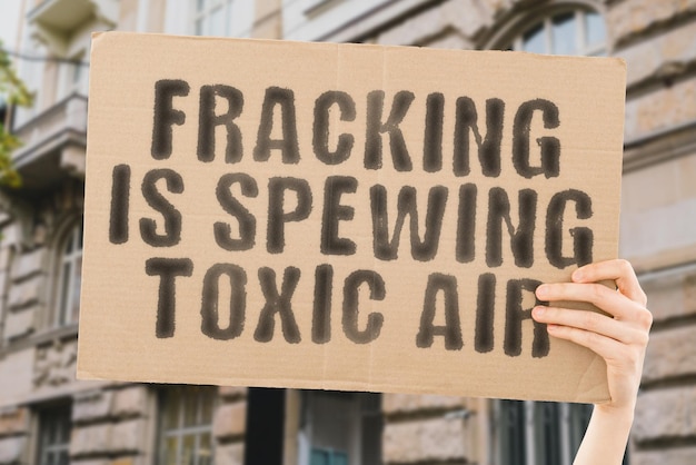 The phrase Fracking Is Spewing Toxic Air on a banner in men's hand with blurred background Disease Issue Dirty Crude Pump Fire Drill Cloud Air Rig Oil Shale Gas Fuel Dusk Pollution