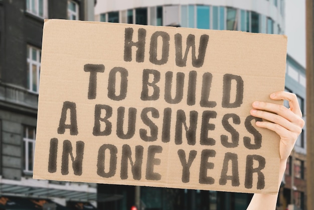 The phrase How to build a business in one year is on a banner in men's hands with blurred background Project Puzzle Revenue Step Built Work Value Target Profit Progress Solution Stack