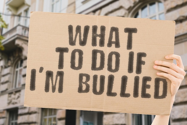 The phrase " What to do if Iâm bullied " on a banner in men's hand with blurred background. Anxiety. Adolescence. Society. Abused. Anger. Friends. Troubled. Stressful. Discrimination