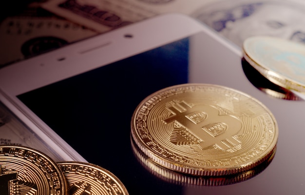 Physical Gold Bitcoin Coin against dollar bills and smartphone on a purple background.