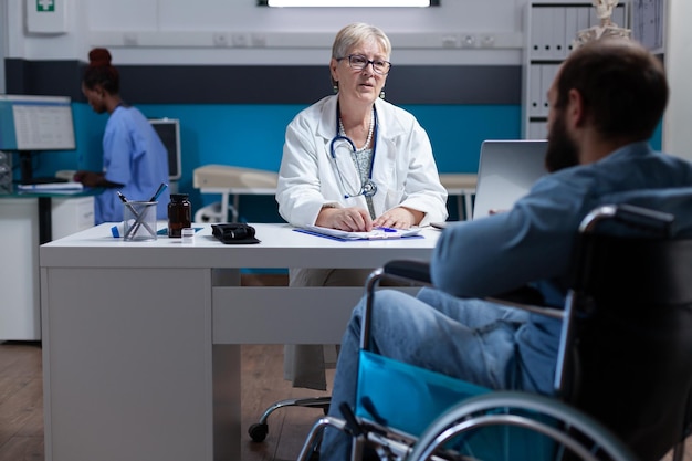 Physician consulting man with disability at checkup visit. General practitioner doing medical examination for healthcare of patient sitting in wheelchair. Woman doctor at consultation.