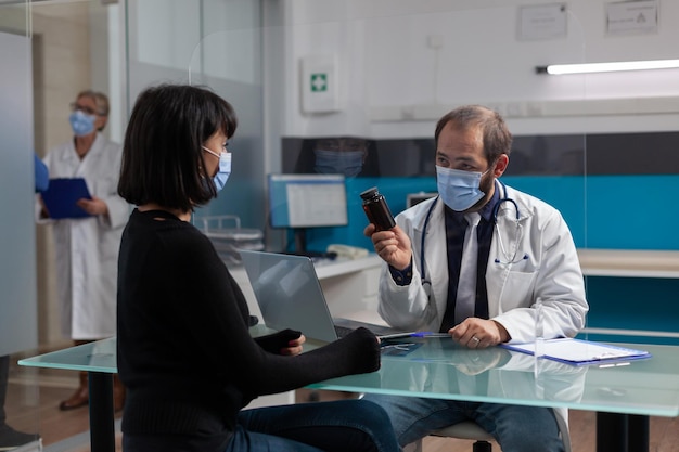 Physician giving bottle of pills to woman for prescription treatment, doing checkup consultation in cabinet. Patient receiving flask of medicaments from medic during covid 19 pandemic.