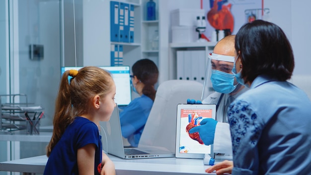 Physician talking about heart functions with patients during coronavirus using tablet. Pediatrician doctor in protection gloves and mask providing clinic medicinal health care services consultation tr