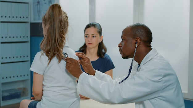 Physician using stethoscope on child to measure heartbeat at checkup visit. General practitioner examining health care of little girl at medical consultation appointment in office with parent.