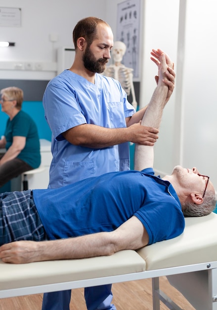 Physiotherapist giving chiropractic massage to elder man, helping with arm muscle recovery. Assistant massaging to stretch hand of old person for kinetotherapy treatment and care.