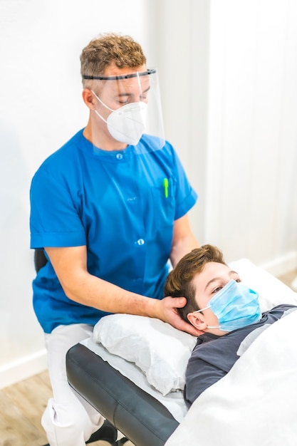 Physiotherapist with mask performing a cranial massage to a child with a mask on the stretcher. Physiotherapy safety measures in the Covid-19 pandemic. Osteopathy, therapeutic chiromassage