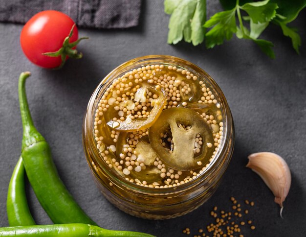 Pickled jalapeno hot pepper in a jar on a dark background with fresh vegetables and herbs The concept of a healthy and delicious homemade snack