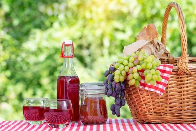 Picnic basket, grapes, juice and jam on a red tablecloth on a natural green background