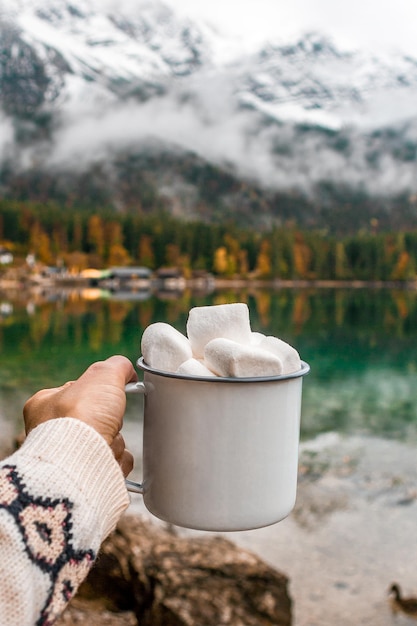 Photo picnic in bavarian mountains germany hand with mug of cacao and marshmallow on the lake background