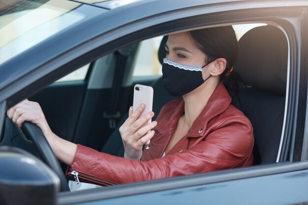 Picture of adorable woman with ponytail holding phone in hands, sanding voice message or setting route black protective mask driving automobile.