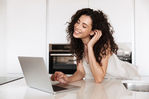 Picture of adorable young woman 20s with curly brown hair wearing silk leisure clothing smiling with perfect teeth, and using silver laptop while leaning on kitchen table