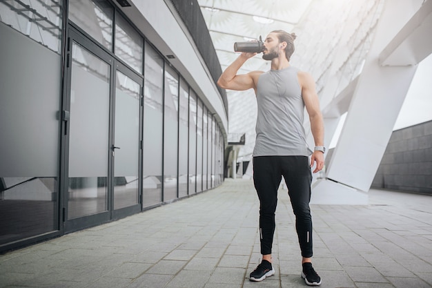 Picture of athletist standing at door and drinking water from black bottle. He has rest. Guy is alone.