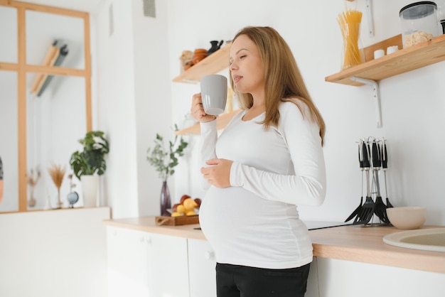 Picture of beautiful casually dressed young brunette female expecting baby, having morning tea in modern stylish kitchen, smiling. Anticipation, expectation, childbearing and motherhood concept