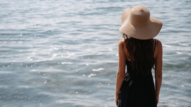 A picture of a beautiful young woman with long dark hair in a dress and a straw hat posing on the beach standing with her back and looking at the endless sea A place for your text