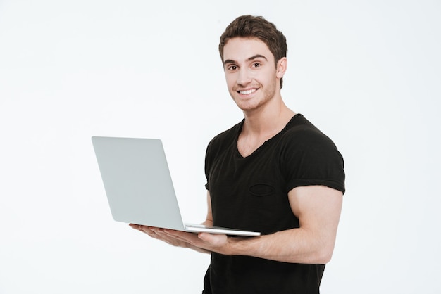 Picture of cheerful young man dressed in black t-shirt standing over white background holding laptop computer and look at camera.