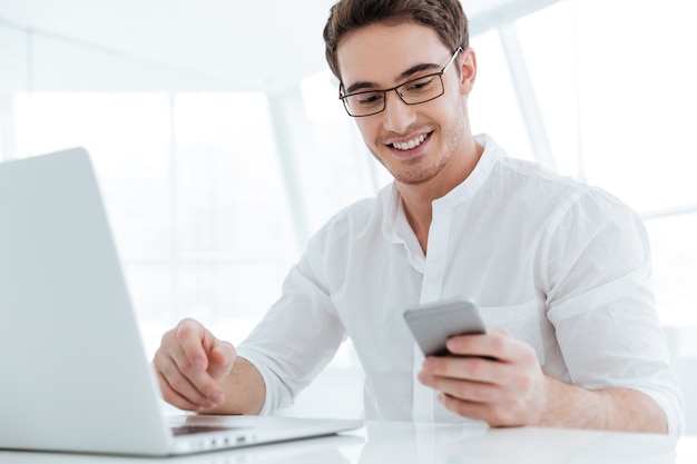 Picture of cheerful young man dressed in white shirt using laptop computer. Look at phone.