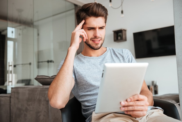 Picture of confused bridal man dressed in grey t-shirt sitting on chair and holding tablet while holding his head with hand