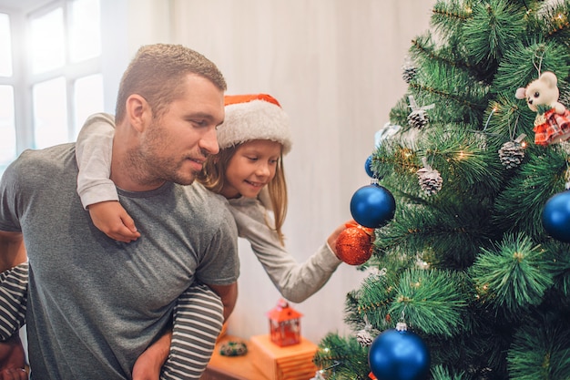 Picture of girl sitting on dad's back and decorating Christmas tree with toys. 