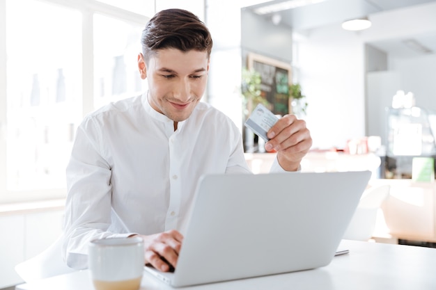 Picture of handsome man dressed in white shirt sitting near cup of coffee while using laptop computer holding credit card in hand. Coworking. Looking at computer.