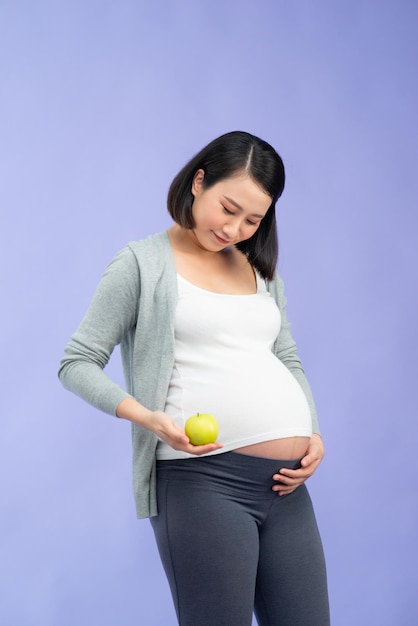 Picture of happy young pregnant asian woman standing isolated over purple background Looking camera holding apple