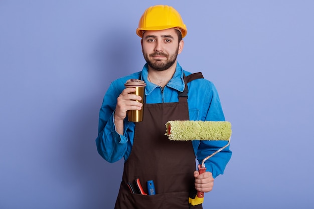 Picture of hard working pleasant repairman holding thermo mug with hot drink and roller for wall painting, having coffee break, wearing uniform with equipment, having rest, enjoying lunch time.