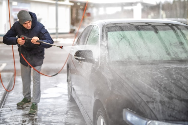 A picture of a man washing a car under high water pressure outdoors in the cold season.