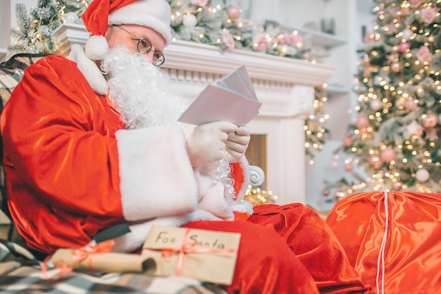 Picture of Santa Claus sitting and reading letters. He holds them in hands. Man is concentrated and serious.