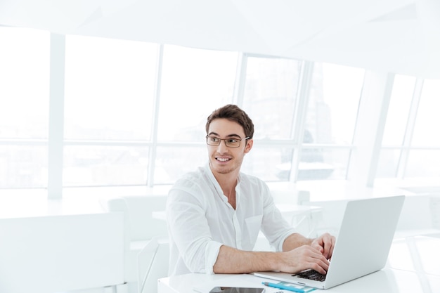 Picture of smiling young man dressed in white shirt using laptop computer. Look aside.