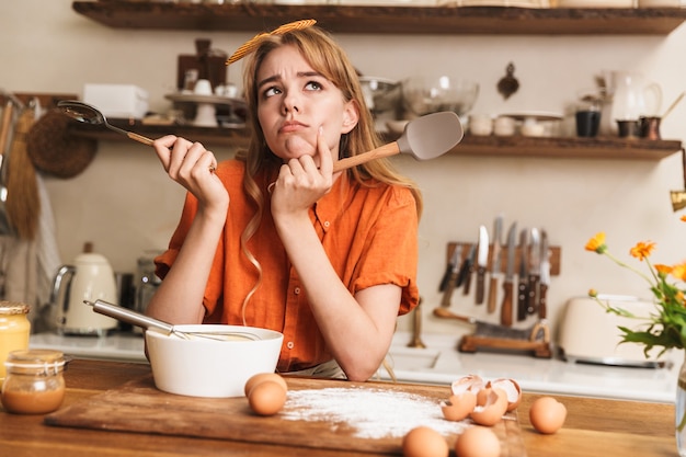 Photo picture of a thoughtful confused sad young blonde girl chef cooking at the kitchen thinking.