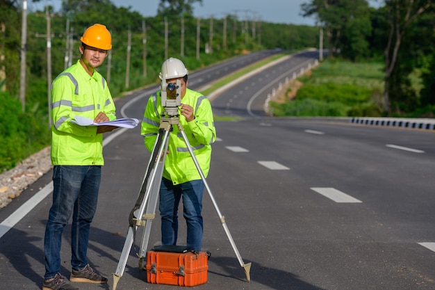 Photo picture of two civil engineers using theodolites measuring land coordinates standing at outdoor theodolites at a road construction site.