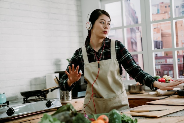 Picture of young amazing emotional woman dancing in kitchen indoors at home listening music with headphones cooking. fresh vegetables putting on wooden counter table. lady in earphones moving body