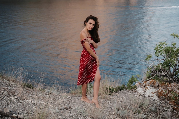 A picture of a young attractive Caucasian woman in a red dress at the edge of a cliff near the beach and the sea