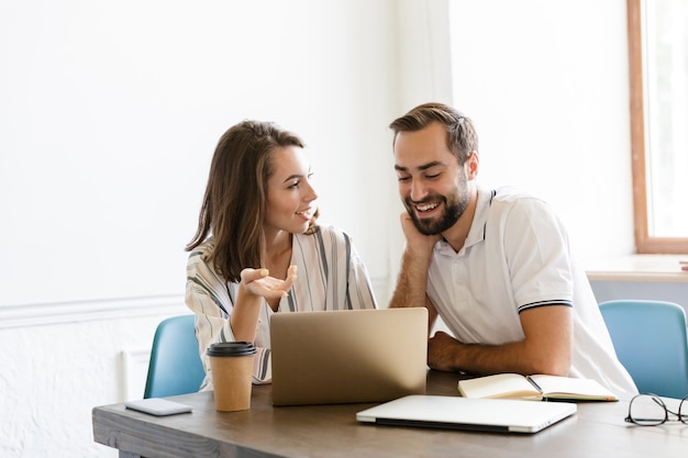 Picture of a young cheerful couple colleagues work with laptop computer indoors in office talking with each other.