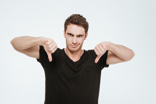 Picture of young handsome man dressed in black t-shirt standing over white background looking at camera and showing thumbs down gesture.