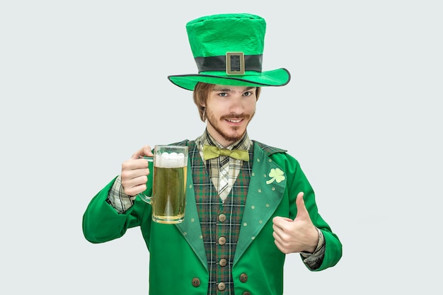 Picture of young happy man in green saint Patrick suit holding mug of beer and smile. He hold big thumb up. Isolated on grey .