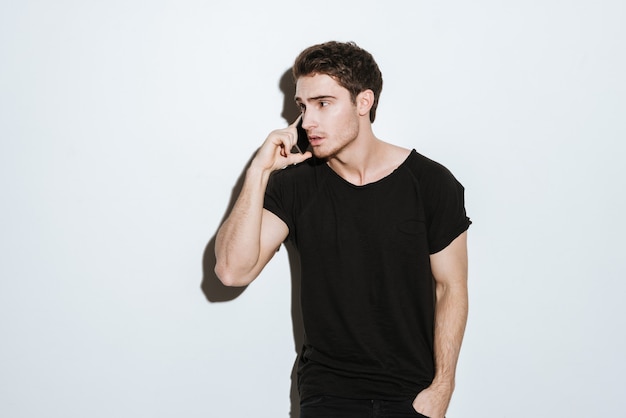 Picture of young man dressed in black t-shirt posing over white background and talking by phone. Looking aside.