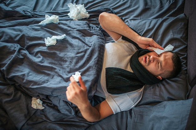 Picture of young man lying on bed and suffering from headache and nose running. He feels bad and exhausted. Young man holds dry napkin on head and shrinking. There are lots of napkins on bed.