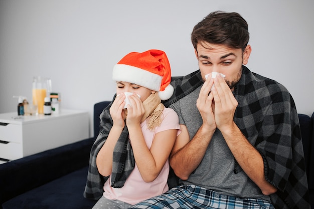 Picture of young man with daughter sitting on sofa and sneezing. They caught cold. Flu and sickness. Celebrating new year of Christmas. Red hat of girl's head. Festive month