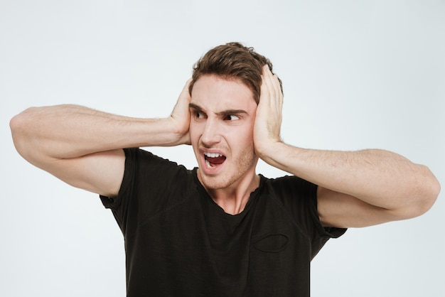 Picture of young screaming man dressed in black t-shirt standing over white background looking aside while holding his head and covering ears.