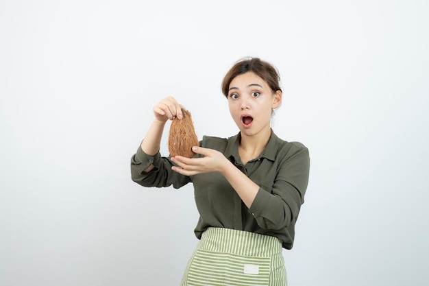 Picture of young woman in apron holding a coconut against white wall . High quality photo