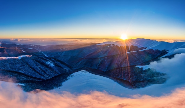 Picturesque winter panorama of mountain hills covered with snow and fir trees on a sunny clear day with the sun and blue sky. Pristine Nature Beauty Concept